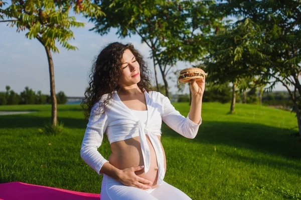 Vacker gravid kvinna med en hamburgare i park — Stockfoto