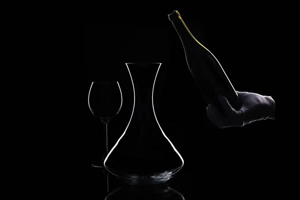 Hand in white glove holds bottle of red wine at bottom and decanter