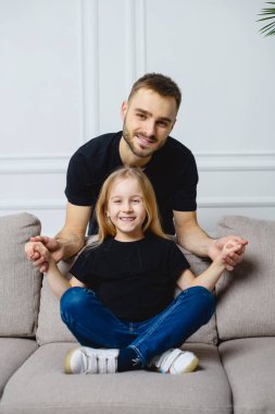 Baba ve kızının kanepedeki portresi. Genç bir kızla genç bir baba. Eğleniyor ve gülümsüyor. Evdeki kanepede benzer bir kıyafetle oturuyor..