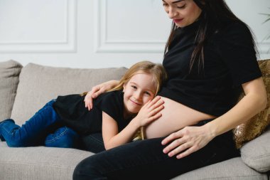 Anne ve kız kanepede oturur ve karnına dokunurlar. İki çocuklu mutlu aile bebek bekliyor. Benzer bir günlük kıyafet - siyah tişört ve kot.