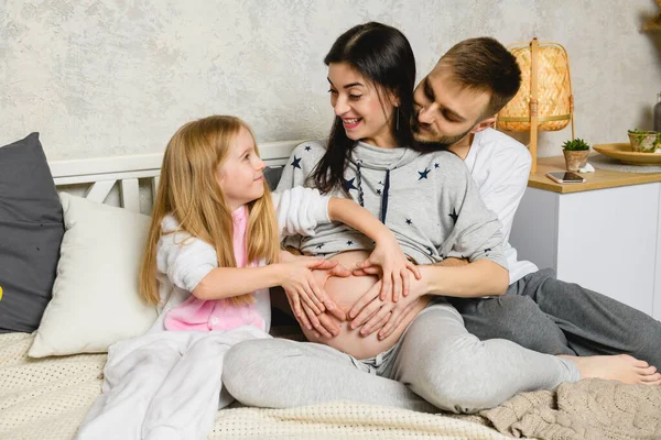 Happy family: daughter and dad hug a pregnant mom, forming a heart from their hands on her belly.