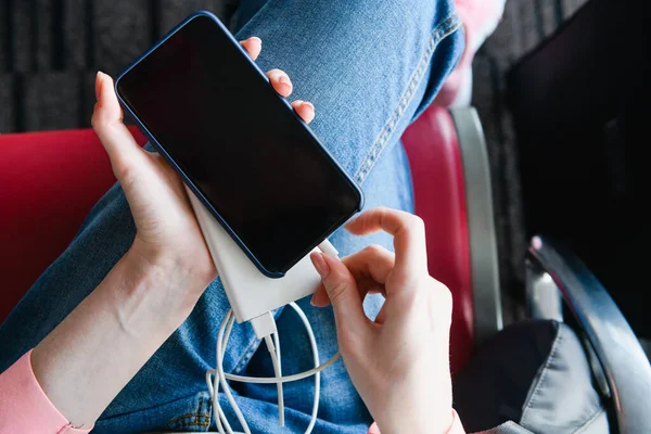 Female in jeans at red airport terminal seat hold portable charger connected to mobile phone to refill battery, up view of black locked screen. Top view.