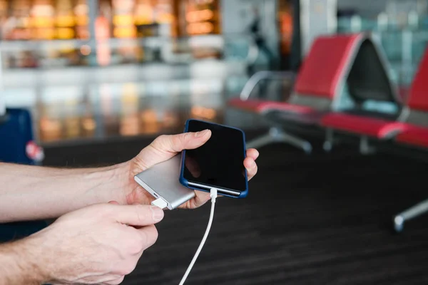 Erkek gezgin, havaalanı arka planında USB tarafından taşınabilir şarj cihazıyla cep telefonunu şarj ediyor. Yakın plan..
