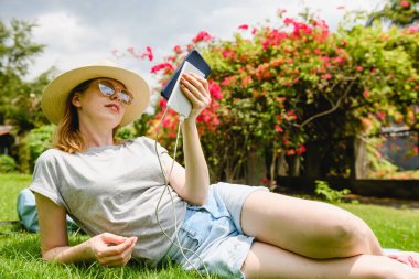 Kadın, parkta piknikte elinde cep telefonu ve taşınabilir güç bankasıyla yeşil çimlerin üzerinde yatıyordu.