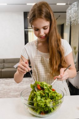 Genç gülümseyen dişi, yeşil salatayı domateslerle karıştırırken iki kaşık tutuyor. Ve sos olarak da yağ, limon suyu, kapalı alanda tuz.