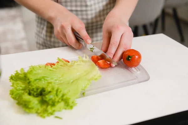 エプロンの女性はナイフを保持し キッチンの背景に健康的なサラダ皿ボウルのためにボード上のトマトと緑をカット — ストック写真