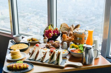 Pencere kenarındaki masada bolca yiyecek ve içecek bulunan otel odası romantik kahvaltısı - kruvasan, meyve, sandviç, ızgara et, çay, meyve suyu, kahve, yumurta