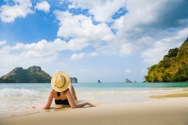 Siyah mayo giyen, beyaz kuma uzanmış, deniz dalgaları ve kayalarla Poda adası manzarasının tadını çıkaran egzotik bir plaj kadını. Arkadan bak..