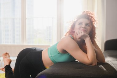 Çekici, güler yüzlü bir kız yurt içi atmosferinde kanepeye uzanmış spor tayt ve yeşil sütyen giyip salgın karantina sırasında evde antrenman yaptıktan sonra rahatlıyor.