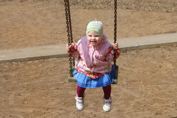 Piccola ragazza carina in cappello oscilla sul parco giochi per bambini con sabbia — Foto Stock