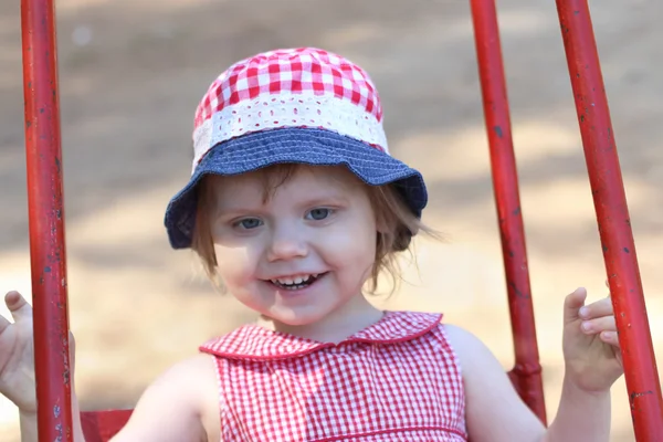 Petite fille heureuse en chapeau balançant sur l'aire de jeux pour enfants au printemps — Photo