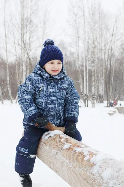 Glad liten pojke sitter på loggen gungbräda i parken under snöfall — Stockfoto