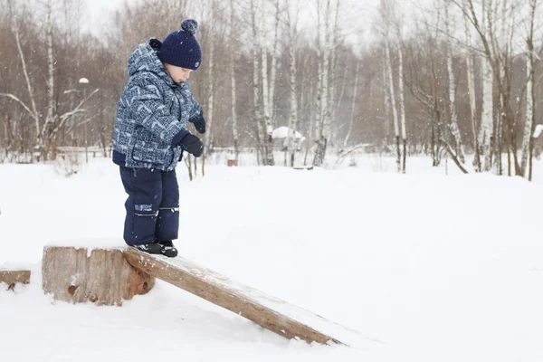 Felice bambino sta sul tronco nel parco durante la nevicata in inverno — Foto Stock