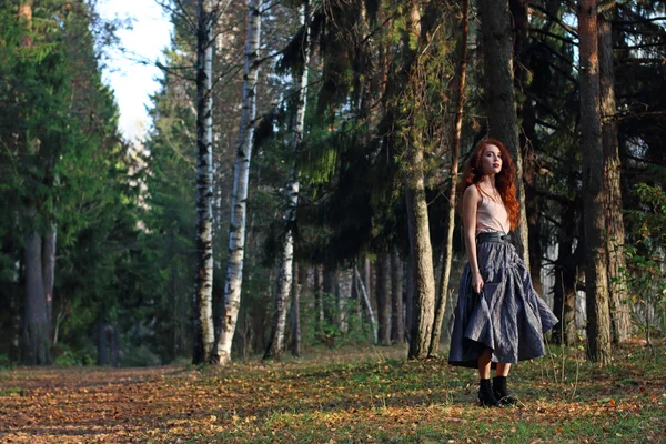 Mooie vrouw staat op droge bladeren in rok in herfst bos — Stockfoto