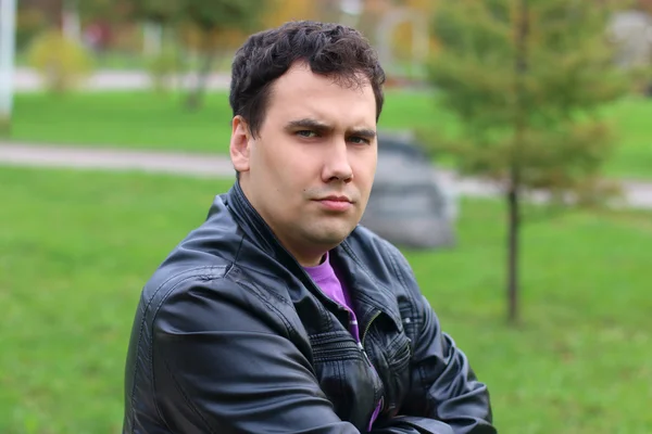 Handsome serious man in leather jacket looks at camera in park — Stock Photo, Image