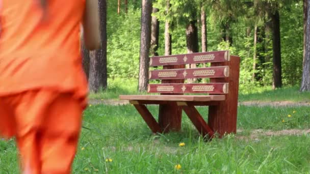 Счастливая маленькая девочка в апельсине сидит на скамейке в зеленом парке — стоковое видео