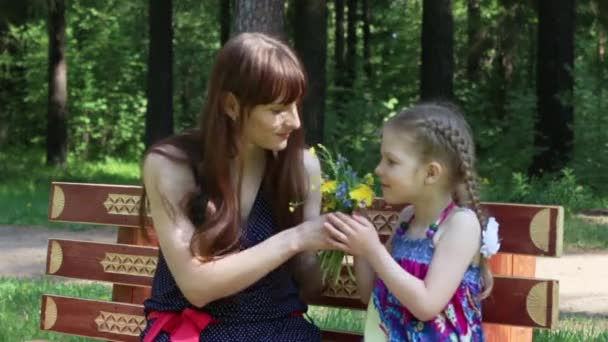 Feliz madre e hija se sientan con flores en el banco en el parque en verano — Vídeo de stock