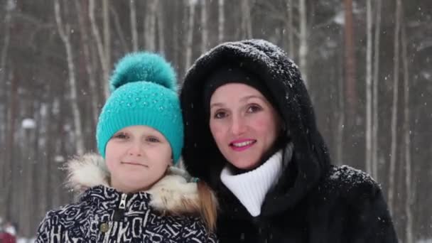 Mother and daughter smile and wave hands during snowfall in winter park — Stock Video
