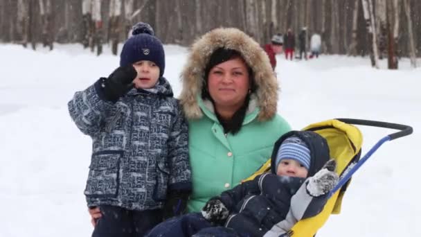 Mother and two sons wave hands, one boy kisses mother during snowfall in winter park — Stock Video