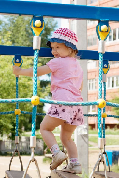 Liten söt flicka i hatt klättrar på lekplats på soliga d — Stockfoto