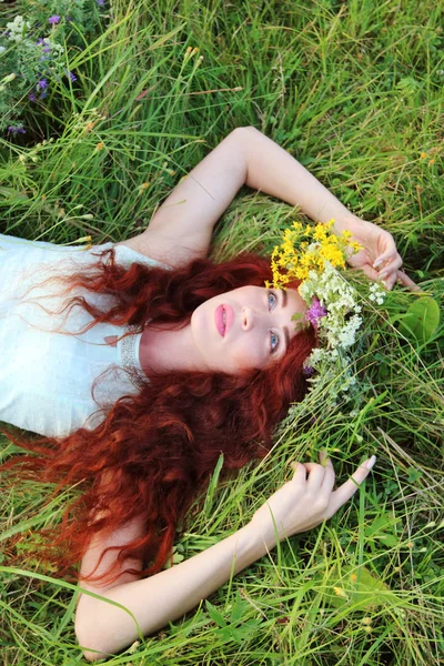 Girl in wreath lies on green grass and looks up on meadow at sum — Stock Photo, Image