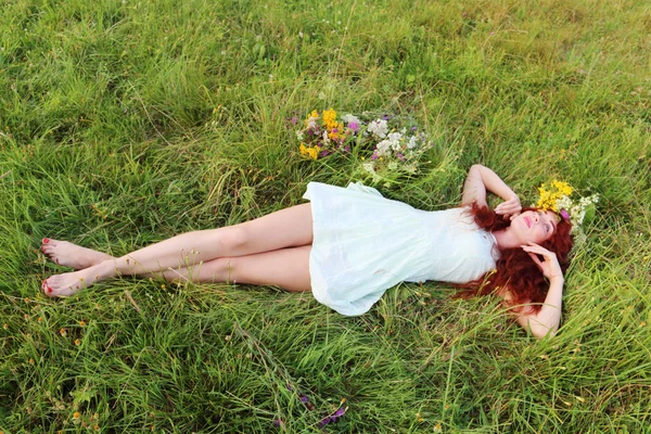 Barefoot girl in wreath lies on grass and looks up on meadow at — Stock Photo, Image