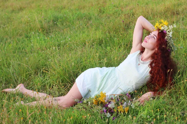 Barefoot woman in wreath lies and looks up in grass on meadow at — Stock Photo, Image