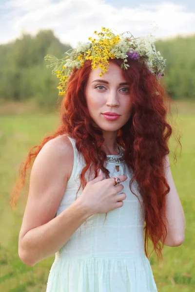 Hezká mladá žena v věnec s červenými vlasy pózy na louce na su — Stock fotografie