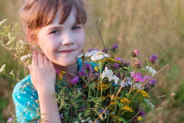 Happy little girl holds wild flowers and smiles in dry field at — Stock Photo, Image