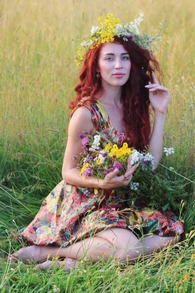 フィールドの草の中に座っている赤い髪と花輪で美しい女性、 — ストック写真