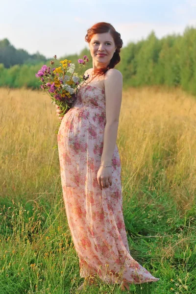 Pretty pregnant woman in long dress poses with wild flowers at s — Stock Photo, Image