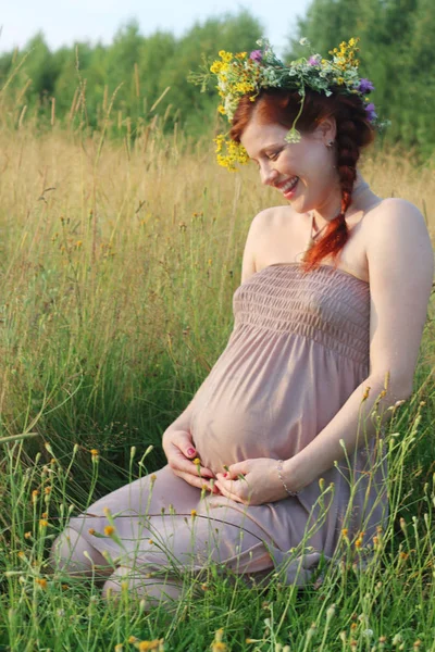 Beautiful pregnant woman in wreath sits and laughs in dry field — Stock Photo, Image