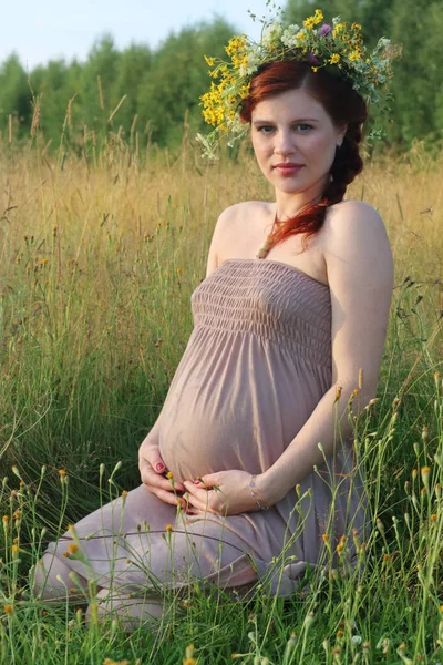 Beautiful freckled pregnant woman in wreath sits in dry field at — Stock Photo, Image