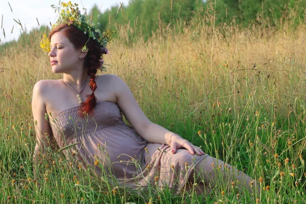 Beautiful pregnant woman in wreath sits and dreams in dry field — Stock Photo, Image