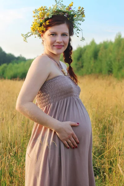 Pretty pregnant woman in wreath poses in dry grass at summer day — Stock Photo, Image