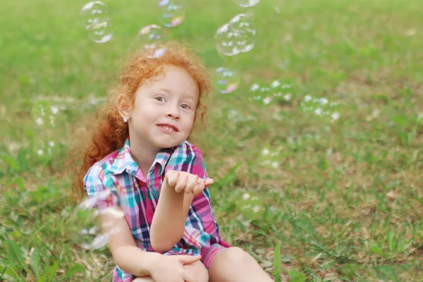 Petite fille aux cheveux roux pose sur l'herbe parmi les bulles en été — Photo