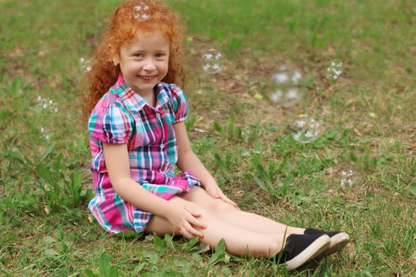 Little girl with red hair sits on grass among bubbles in summer — Stock Photo, Image