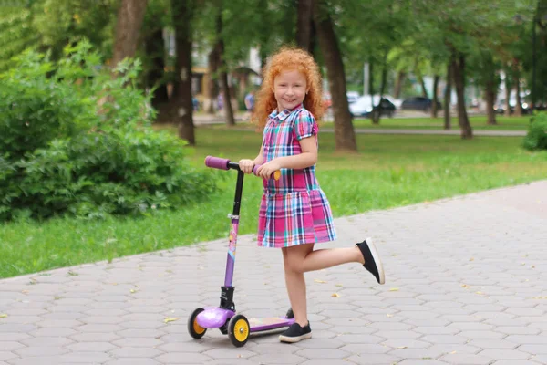 Happy little girl with red hair smiles with scooter in summer pa — Stock Photo, Image