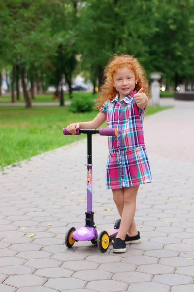 Happy little girl with red hair thumbs up with scooter in summer — Stock Photo, Image
