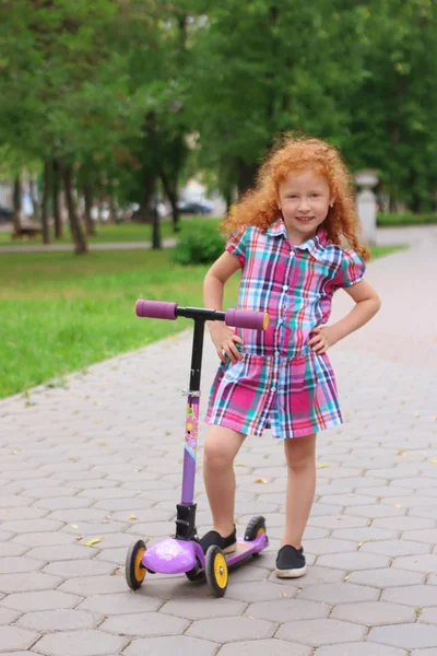 Happy little girl with red hair stands with scooter in summer pa — Stock Photo, Image