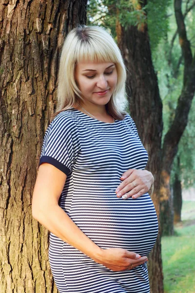 Happy beautiful pregnant woman in dress poses near tree in sunny — Stock Photo, Image