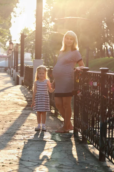 Pregnant mother and her little daughter pose near railing in sun — Stock Photo, Image