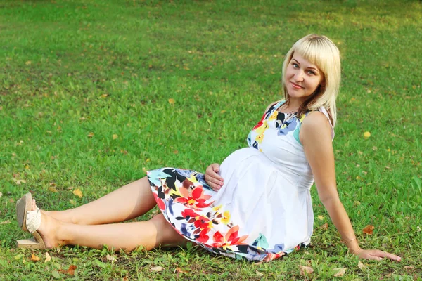 Feliz hermosa mujer embarazada en vestido se sienta en la hierba verde en s —  Fotos de Stock