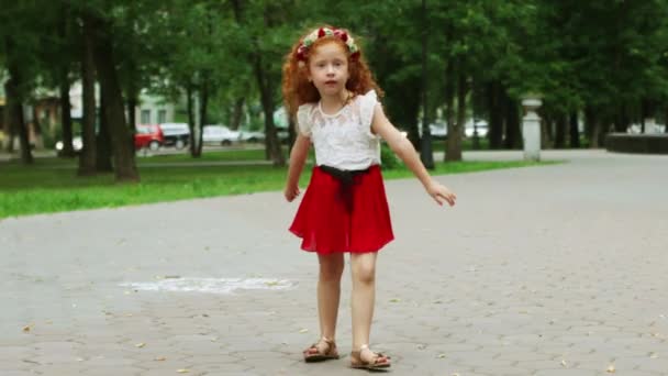 Маленькая девочка с рыжими волосами танцует в зеленом летнем парке — стоковое видео