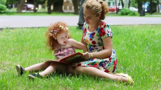 Kis lány és az anyja olvassa el a könyvet, a gyep a parkban nyári — Stock videók