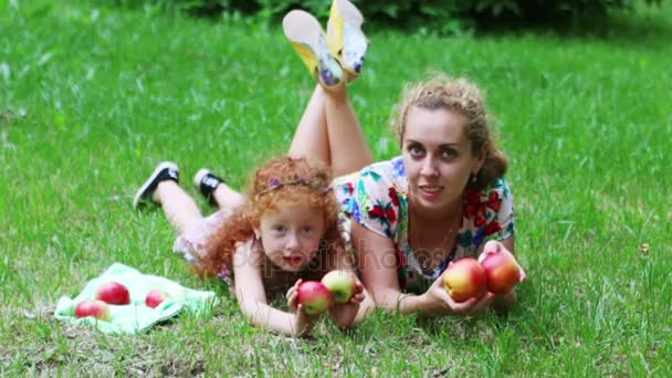 Маленькая девочка с рыжими волосами и ее мать играют с яблоками на газоне в зеленом летнем парке — стоковое видео