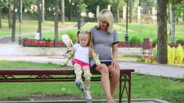 日当たりの良い公園のベンチに座っている妊娠中の女性とローラー スケートを持つ少女 — ストック動画