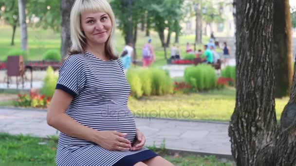 Zwangere vrouw zit op bankje, glimlacht en haar buik lijnen in zonnig park — Stockvideo