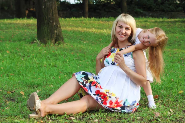 Happy little girl and pregnant mother pose on green grass in sun — Stock Photo, Image