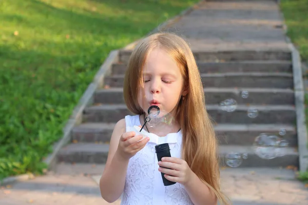 Little cute girl blows bubbles near stairs in green sunny park — Stock Photo, Image